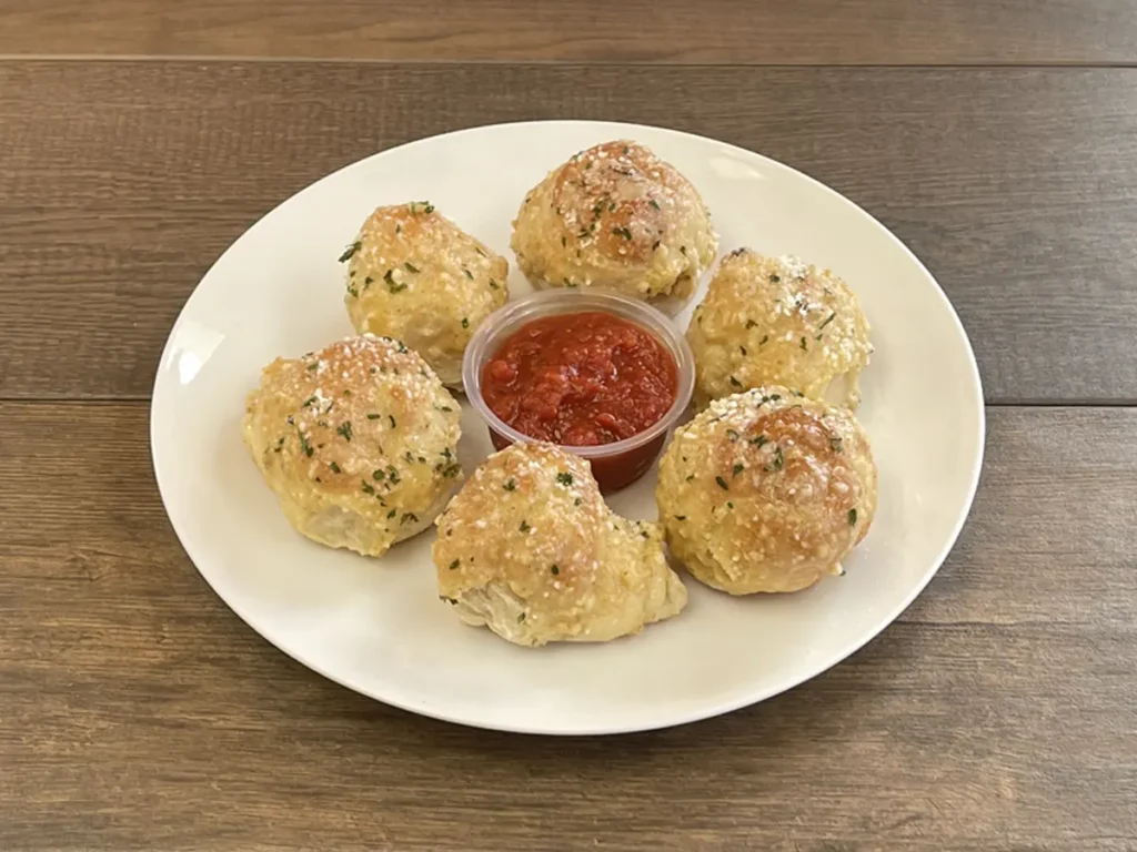Six garlic knots with garlic spread and herbs around a cup of marinara